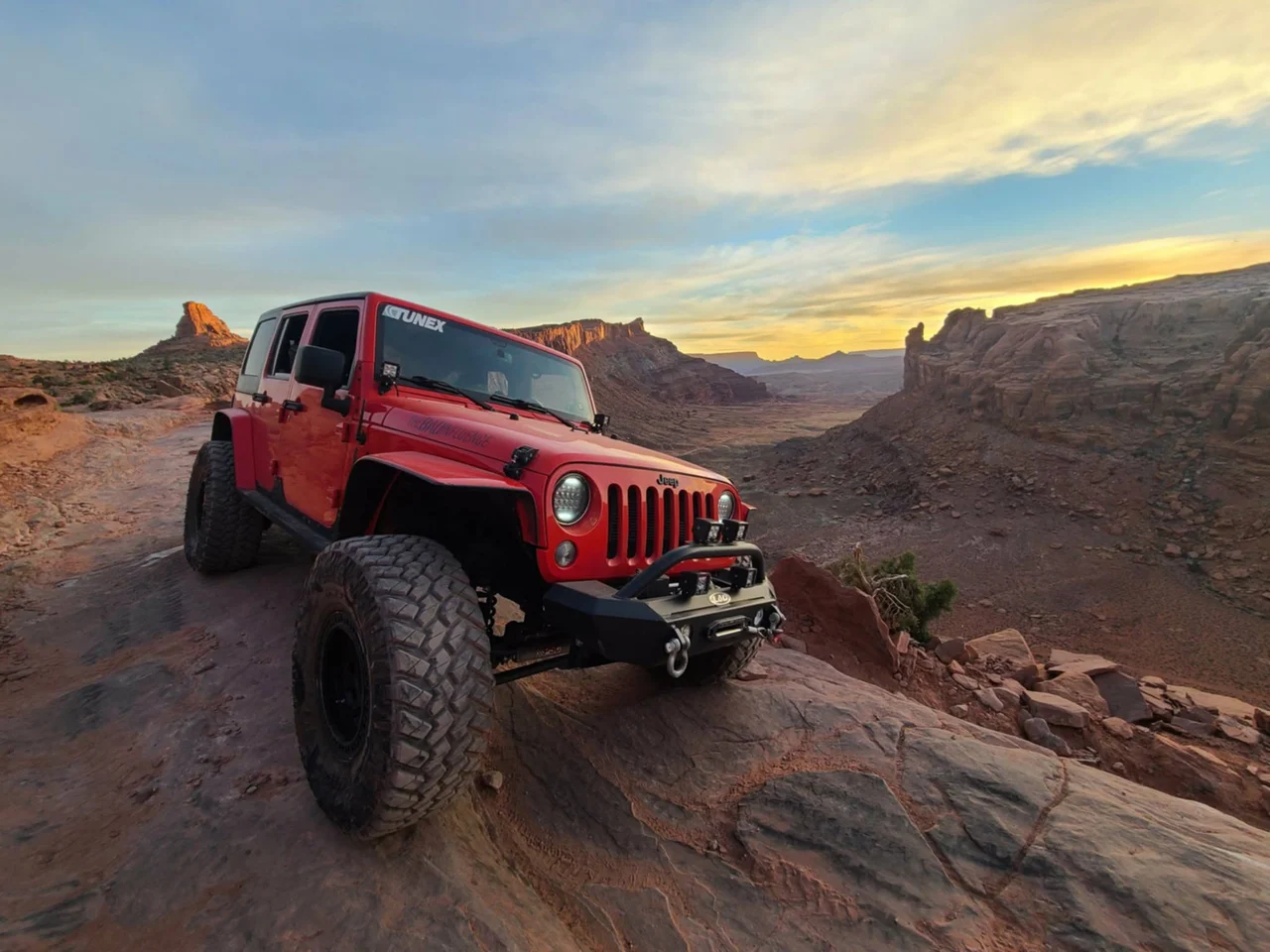 Red Jeep on a rocky trail overlooking red-rock canyons beneath a golden sky